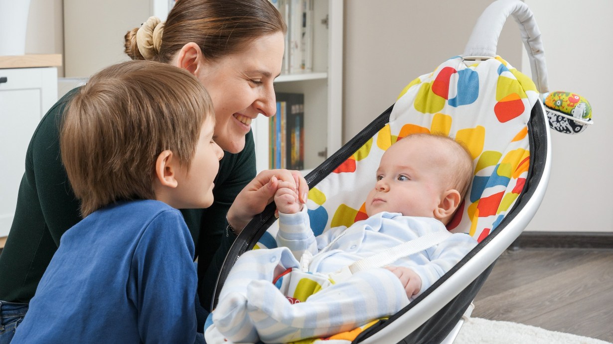 A woman and child smile at an alert baby in a rocker.
